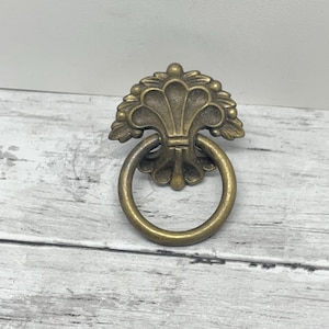 May include: Antique brass drawer pull with a decorative floral design and a round ring handle. The hardware features a detailed, ornate pattern and a warm, aged finish. Ideal for vintage furniture restoration or adding a classic touch to cabinets.