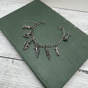 May include: A silver charm bracelet featuring various charms, including a race car, a skier, a diver, a golfer, an archer, and a dancer. The bracelet is displayed on a green book.