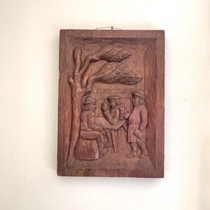 May include: A rectangular brown wooden wall plaque featuring a relief carving of three figures gathered under a tree. Two figures are seated at a table while one stands nearby, all depicted in period clothing. A metal wire is attached for hanging.