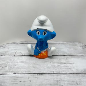 May include: A blue Smurf figurine with a white hat and orange shorts. The Smurf is sitting with its legs spread. The figurine has black eyes and is on a white and gray wooden surface.