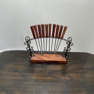 May include: A vintage fondue fork set with a dark wood base and handles. The set includes a black metal frame with decorative scrollwork. The forks are arranged in a fan shape, perfect for entertaining.