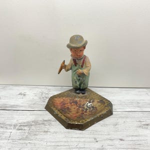 May include: A vintage figurine of a young boy in overalls and a hat, holding a carrot. The figurine is painted in muted colors and stands on a diamond-shaped base with a textured surface. The base is approximately 3 inches wide.