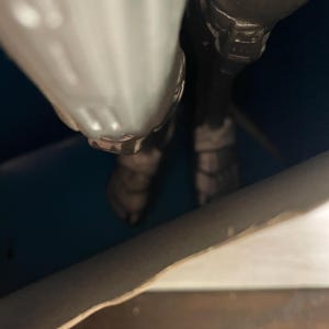 May include: Close-up of a metallic gray action figure. The figure's legs and lower torso are visible, with detailed armor and boots. The figure is positioned against a dark blue background.