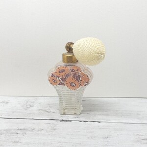 May include: A clear glass perfume bottle with a textured, basket-weave base and a floral decal featuring orange and brown flowers. The bottle has a gold-toned metal collar and a round, cream-colored fabric atomizer bulb attached to the top.