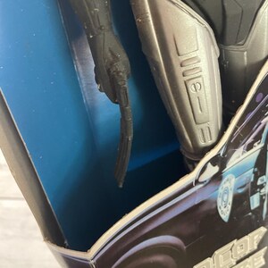 May include: Close-up of a Robocop action figure in its original packaging. The figure is black and silver, with a detailed design. The packaging features the words "RoboCop Figure" in a stylized font, with a police car graphic.