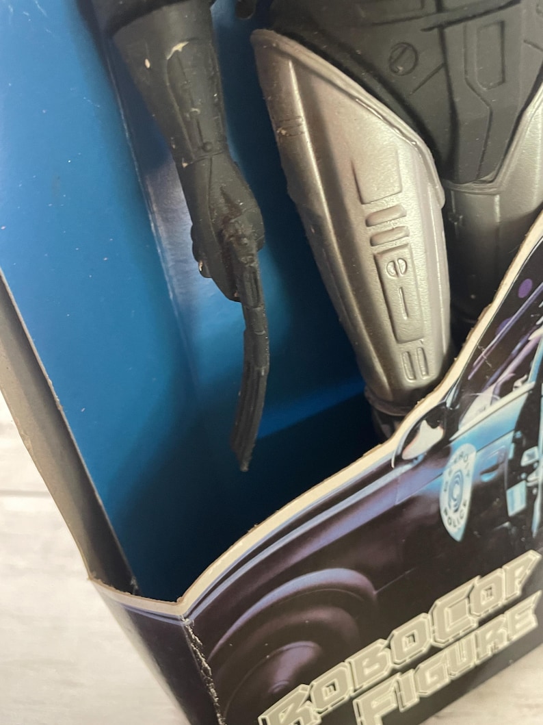 May include: Close-up of a Robocop action figure in its original packaging. The figure is black and silver, with a detailed design. The packaging features the words "RoboCop Figure" in a stylized font, with a police car graphic.