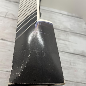 May include: A black box with a white and gray striped design on the side. The box has some wear and tear, with visible scratches and scuffs. The box is on a light gray wooden surface.