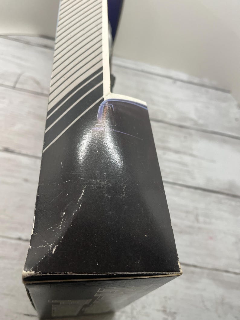 May include: A black box with a white and gray striped design on the side. The box has some wear and tear, with visible scratches and scuffs. The box is on a light gray wooden surface.