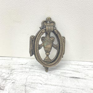 May include: Antique brass-toned door knocker with an oval frame and a central urn design. The knocker features ornate detailing and a small loop at the top for hanging. The overall design is symmetrical and classic.