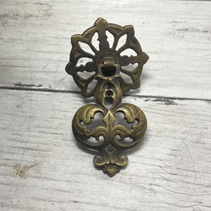 May include: An antique brass drawer pull with a floral design at the top and a decorative, oval-shaped bottom. The hardware is made of brass and has an aged, vintage appearance. The drawer pull is designed for furniture restoration.
