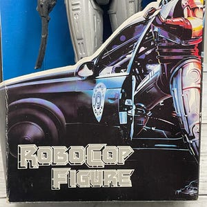 May include: A RoboCop action figure in its original packaging. The box features a graphic of RoboCop standing next to a police car. The words "RoboCop Figure" are printed in silver block letters on a black background.