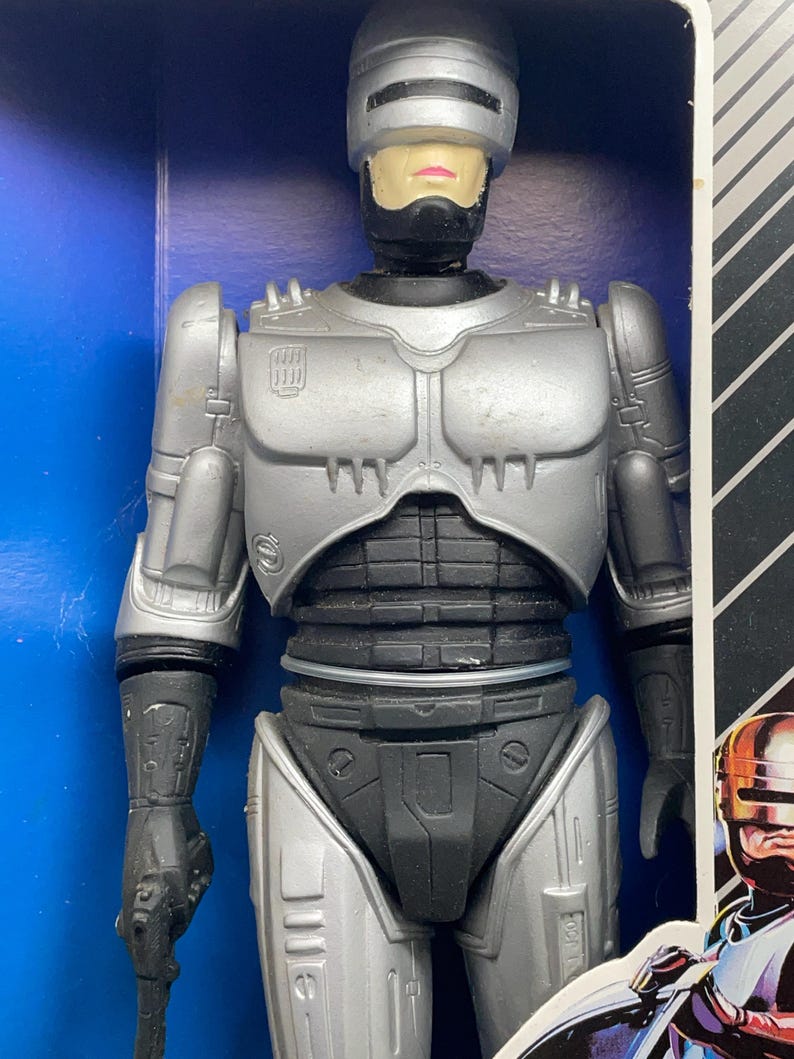 May include: A silver and black Robocop action figure in its original packaging. The figure has a helmet, chest armor, and black accents. The packaging is blue with black and white stripes.