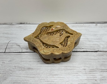 May include: A beige, carved stone box with a floral design. The box has a lid with intricate cut-out patterns and a raised floral motif. The box is octagonal in shape and sits on a white and gray wooden surface.