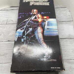 May include: A Robocop action figure box. The black box features the words "ROBOCOP FIGURE" in silver lettering. The image on the box shows Robocop standing next to a police car. The box is marked "MADE IN CHINA".