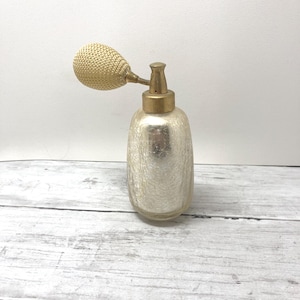 May include: A vintage-style perfume atomizer featuring a cylindrical glass bottle with a crackled, mercury-glass finish. The bottle is topped with a metallic gold-toned collar and a matching gold-toned bulb pump covered in a textured, woven mesh.