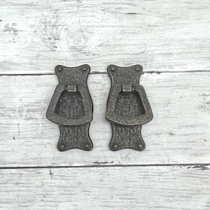 May include: Two antique bronze-colored drawer pulls. Each pull has a rectangular handle with a decorative, textured design. The pulls are mounted on a light-colored wooden surface.
