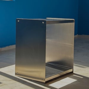 Op de afbeelding: Een rechthoekige opbergeenheid met open voorzijde, vervaardigd uit geborsteld roestvrij staal. De structuur heeft vlakke zijpanelen die op de hoeken zijn vastgezet met zichtbare metalen bevestigingen, wat een hol, doosachtig frame vormt.