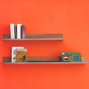 Könnte beinhalten: Zwei horizontale Metallregale an einer leuchtend orangefarbenen Wand. Das obere Regal enthält drei Bücher. Das untere Regal enthält zwei Bücher und ein kleines rechteckiges Dekoschild mit einem Schulbus-Motiv und dem Text PA SCHOOL BUS 64.