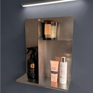 Peut inclure: Une étagère murale en acier inoxydable brossé à deux niveaux. L'étagère supérieure contient une bougie en verre avec un couvercle en liège. L'étagère inférieure contient un vaporisateur noir, un tube de BB crème pêche et un flacon blanc à bouchon argenté.