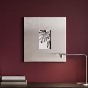 Puede incluir: Un marco de pared cuadrado de metal plateado cepillado con un recorte rectangular central que muestra una impresión botánica en blanco y negro de dos hojas de monstera. El marco tiene un acabado metálico liso y un diseño geométrico.
