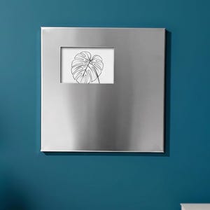 Puede incluir: Un marco de fotos cuadrado de metal plateado cepillado con una superficie lisa y reflectante. El marco presenta un recorte rectangular en el cuadrante superior izquierdo, que muestra una inserción blanca con un dibujo lineal negro de una sola hoja de monstera.