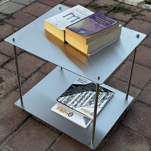 May include: A two-tiered metal side table with a brushed silver finish, featuring four thin cylindrical legs and flat square shelves secured by small rounded bolts. Two books rest on the top shelf, and two thin brochures are on the bottom shelf.