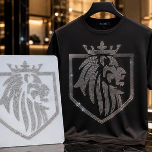 May include: A black short-sleeved t-shirt featuring a large, silver-toned rhinestone lion head emblem inside a shield topped with a crown. A matching rhinestone lion emblem on a white square board is positioned to the left of the t-shirt.