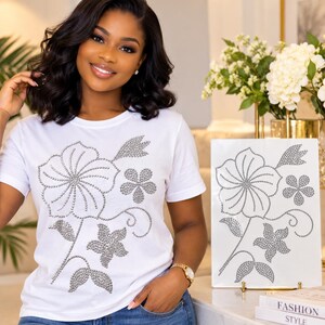 May include: A white short-sleeved t-shirt featuring a large floral design composed of silver rhinestones. The graphic depicts a central large flower with smaller surrounding blossoms, leaves, and swirling stems, all rendered in sparkling silver studs.