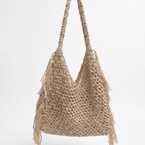 Può includere: Una borsa a spalla all'uncinetto color beige caratterizzata da un motivo a rete a maglia aperta, lunghe frange sui lati e piccole paillettes metalliche integrate nel filato. La borsa include un unico spallaccio integrato.