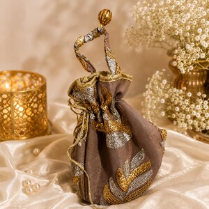 Könnte beinhalten: Ein taupefarbener Beutel mit Kordelzug, der mit aufwendigen goldenen und silbernen Paillettenstickereien in Blattform verziert ist. Die Tasche verfügt über einen passenden Henkel mit Pailletten und eine kleine, runde, strukturierte Goldperle.