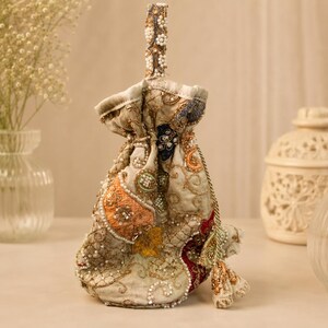 May include: A drawstring pouch bag featuring intricate embroidery in shades of gold, orange, and deep red, embellished with white beads and sequins. The bag has a structured handle and a matching beaded tassel hanging from the side drawstring closure.