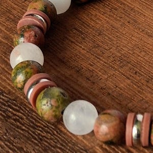 Op de afbeelding: Een kralenarmband met afwisselend ronde groen en roze gevlekte stenen kralen, doorschijnende witte bolvormige kralen en platte, cirkelvormige mauvekleurige tussenstukjes, geaccentueerd met dunne, reflecterende zilverkleurige metalen ringen.