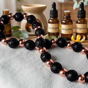 Puede incluir: Una pulsera de cuentas que alterna cuentas redondas de piedra de lava negra porosa y cuentas metálicas lisas de color cobre, dispuestas en un patrón circular continuo sobre una superficie de tela blanca texturizada.