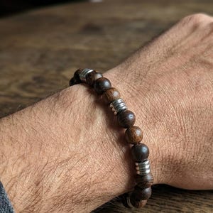 May include: A beaded bracelet featuring alternating dark brown wooden spheres and polished silver-toned metal spacers. The round beads vary in shade from light to dark brown, creating a natural texture, while the smooth metallic accents are placed at intervals along the circular elastic band.