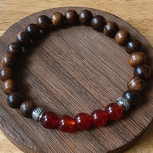 May include: A stretch bracelet featuring dark brown, wood-grain textured spherical beads and five translucent, deep red spherical beads. The red beads are separated from the wooden beads by two small, silver-toned metal spacers with a textured finish.