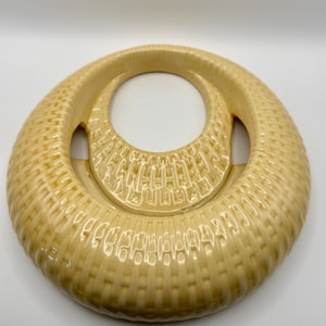 May include: A circular, cream-colored ceramic object with a glossy finish and a textured, woven-like pattern across its surface. The piece features a large, central open space and a smaller, crescent-shaped cutout, creating a distinct, hollow, ring-like structure.