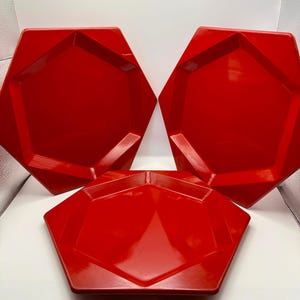 May include: Three glossy, bright red plates with a distinct hexagonal shape. Each plate features a flat central surface surrounded by angled, geometric edges that create a faceted appearance. The plates are arranged in a triangular formation.