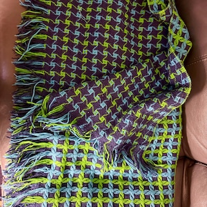 May include: A woven throw blanket featuring a repeating geometric pattern in deep purple, lime green, and light blue. The textile has a textured surface with a fringe border along the edges, showcasing a consistent grid-like design throughout.