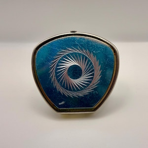 May include: A triangular compact mirror with a metallic silver frame and a vibrant blue enamel top surface. The center of the blue panel features a circular, starburst-like engraved pattern with fine, radiating lines that create a sense of motion.