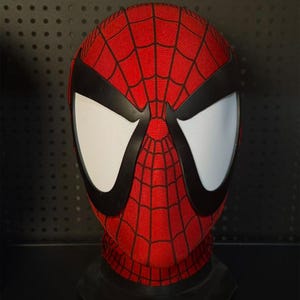 May include: A red fabric mask featuring a black web pattern across the surface. The mask includes large, white, mesh-covered eye lenses framed by thick black borders, designed to cover the entire head and neck area for a full-face costume look.