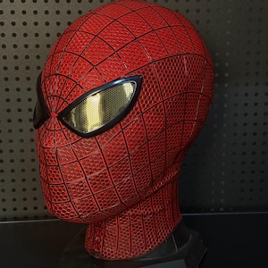 May include: A red full-head mask featuring a raised black web pattern across the entire surface. The mask includes two large, almond-shaped eye lenses with a metallic gold mesh finish, framed by thick black borders, creating a distinct superhero look.
