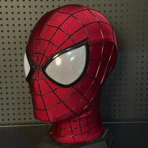 May include: A red full-head mask featuring a textured fabric surface with a black web pattern overlay. The mask includes two large, white, mesh-covered eye lenses framed in black, designed to cover the entire head and neck area completely.