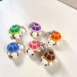 May include: Six small, handmade crochet turtle keychains, each featuring a white head and limbs with black bead eyes. The shells are crafted in distinct colors: green, purple, orange, blue, pink, and brown, all attached to silver metal key rings.