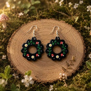 Puede incluir: Un par de pendientes circulares negros con borde festoneado, que presentan un diseño pintado a mano de pequeñas flores blancas, enredaderas verdes y setas rojas con puntos blancos, colgados de ganchos de metal de tono plateado.