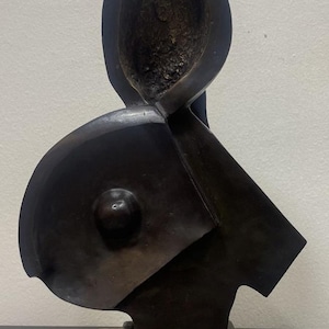 May include: A dark bronze-toned abstract sculpture featuring a smooth, curved form with a central protrusion and a textured, hollowed-out upper section. The sculpture is mounted on a rectangular, speckled light gray and beige stone base.