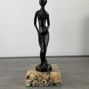 May include: A dark, smooth, abstract sculpture of a standing figure with elongated limbs and a rounded head. The figure is mounted on a square, polished base featuring a speckled, multi-colored granite pattern with tones of beige, black, and gray.