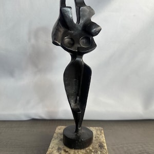 May include: A dark brown, abstract metal sculpture featuring a slender, elongated form with two circular indentations near the top, mounted on a square, speckled beige and grey stone base with a thin red felt layer along the bottom edge.