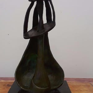 May include: A dark bronze-colored abstract sculpture featuring a tall, slender central form topped with a small, flat, horizontal element. The sculpture has a wide, hollow, bell-shaped base and is mounted on a square, dark, polished stone pedestal.