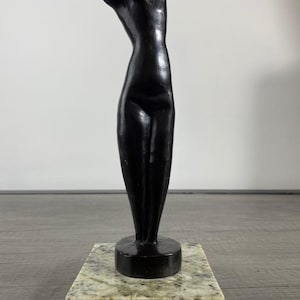 May include: A smooth, black, abstract sculpture of a standing human torso with a curved silhouette. The figure is mounted on a square, polished, light-colored granite base with speckled gray and beige patterns, creating a stark contrast.