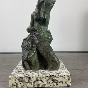 May include: A dark green bronze sculpture depicting an abstract human figure with elongated features and a textured surface. The sculpture is mounted on a rectangular base made of speckled white and gray granite with a polished finish.